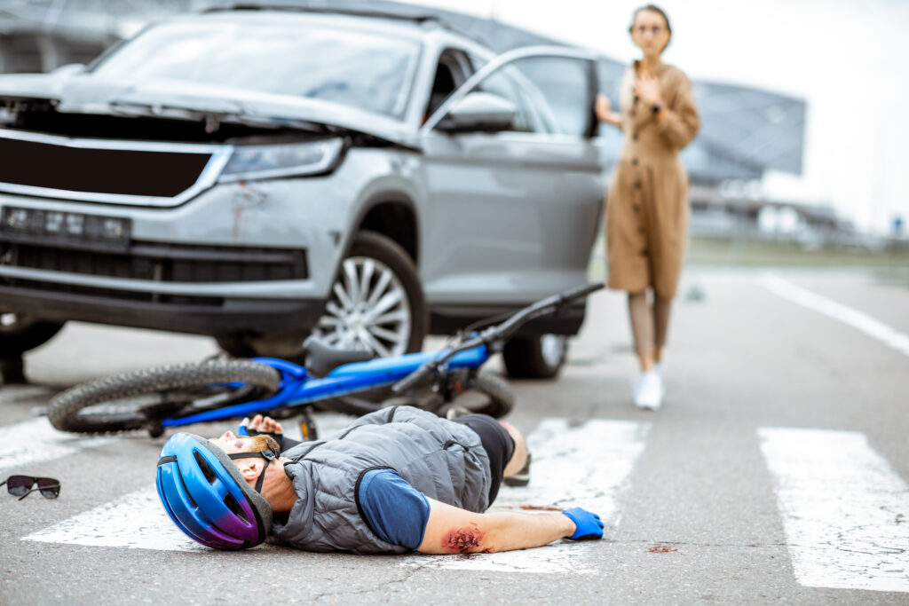 Road sideswipe accident with injured cyclist lying on the pedestrian crossing near the broken bicycle and worried woman driver and car on the background