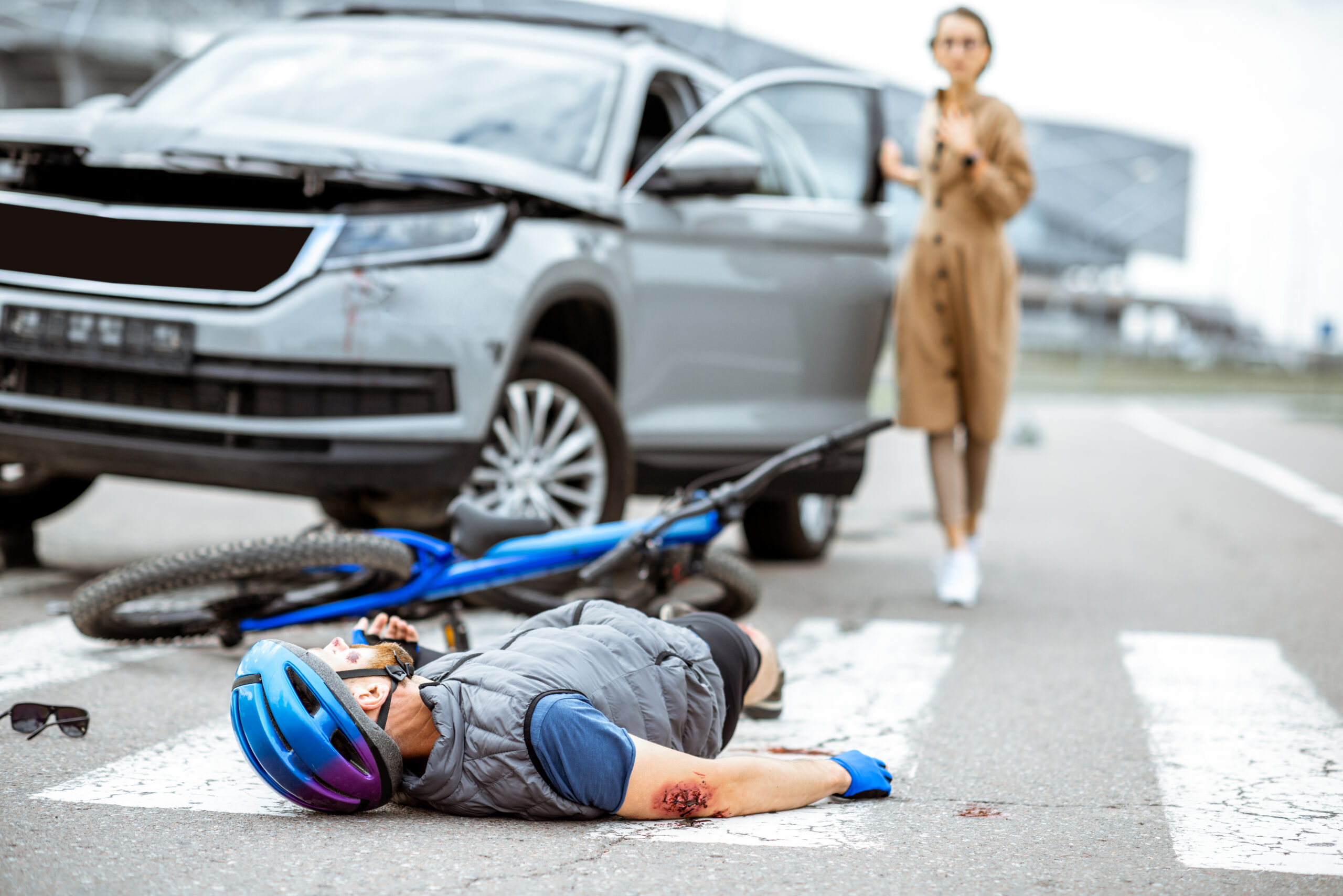 Road sideswipe accident with injured cyclist lying on the pedestrian crossing near the broken bicycle and worried woman driver and car on the background